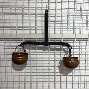 Può includere: Una bilancia in stile vintage con un telaio in metallo nero e due piccole ciotole di rame sospese ai bracci. La bilancia è appesa su uno sfondo a griglia bianca, mostrando il suo design simmetrico e l'estetica invecchiata.