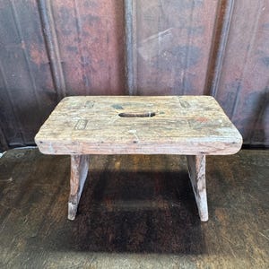 May include: A rectangular wooden stool with a weathered, light brown finish and visible grain. The top surface features a central rectangular cutout handle and visible joinery details at the corners. The stool stands on two sturdy, angled legs.