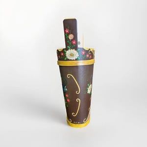 Puede incluir: Un recipiente de madera marrón con un diseño floral decorativo. El recipiente tiene un borde y una base amarillos, con un asa en la parte superior. El diseño floral incluye flores blancas y rojas.