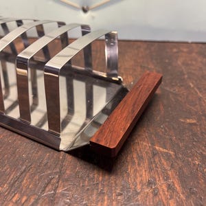 Pode incluir: Um porta-torradas em aço inoxidável com uma pega em madeira escura. O suporte tem várias ranhuras para segurar fatias de torrada. A pega é retangular e presa à base do suporte. O suporte está sobre uma superfície de madeira escura.