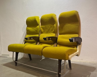 Könnte beinhalten: Ein Satz von drei gelben Vintage-Flugzeugsitzen. Die Sitze haben ein Fischgrätenmuster und Holzarmlehnen. Die Beine sind dunkelbraun mit einer silbernen Stange. Die Sitze stehen vor einer weißen Wand.