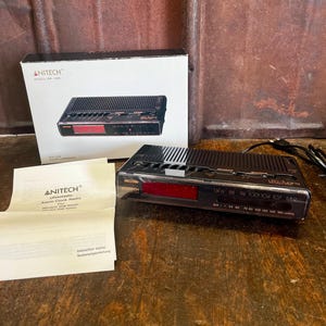 Peut inclure: Un radio-réveil noir Anitech avec un affichage numérique rouge, un cordon d'alimentation noir, une boîte blanche illustrée du produit et un manuel d'instructions blanc plié portant le texte Anitech Uhrenradio Alarm Clock Radio.