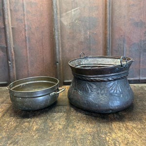 Peut inclure: Deux pots en métal antiques. L'un est un petit bol avec deux poignées. L'autre est un pot plus grand et orné avec une bande décorative en forme de corde autour du haut et des motifs floraux en relief. Les deux sont de couleur foncée.