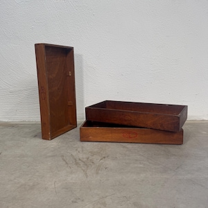 Könnte beinhalten: Drei rechteckige Holztabletts mit mittelbraunem Finish. Ein Tablett steht vertikal auf der Seite, während die anderen beiden horizontal gestapelt sind. Jedes Tablett weist auf der Vorderseite ein kleines, aufgedrucktes rotes Grafiklogo auf.