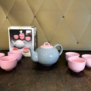 May include: A grey teapot with a pink lid and six pink teacups. The teapot and teacups are arranged on a wooden surface.