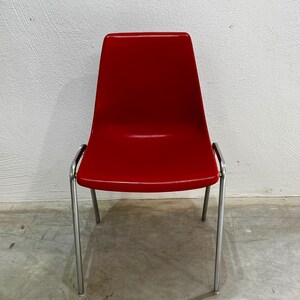 Può includere: Una sedia rossa con finitura lucida. La sedia ha uno schienale curvo e una seduta sostenuta da quattro gambe cromate. La sedia è di un colore rosso pieno e vibrante, e le gambe sono sottili e metalliche.