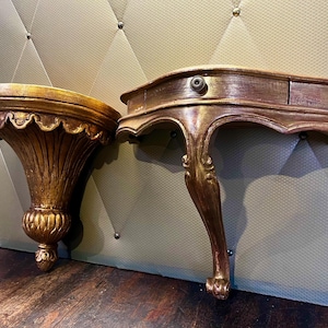 Peut inclure: Deux étagères murales de style antique avec une finition dorée. L'une est une étagère à festons, l'autre est une petite table avec un tiroir et des pieds incurvés. Les étagères sont en bois et présentent un design orné.