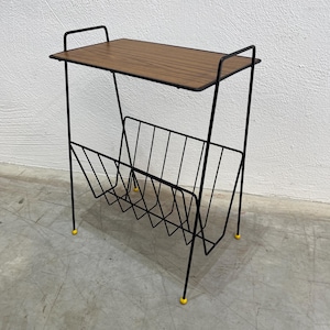Puede incluir: Una mesa auxiliar y revistero de estilo moderno de mediados de siglo. La mesa tiene una tapa rectangular con vetas de madera y un marco de metal negro. El revistero es de alambre negro y tiene patas amarillas.