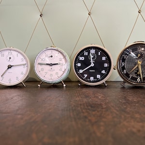 Può includere: Tre sveglie vintage con quadranti di colori diversi. Gli orologi sono bianchi, verdi e neri con numeri bianchi. Gli orologi sono tutti impostati su orari diversi.