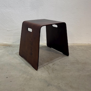 Könnte beinhalten: Ein dunkelbrauner Hocker aus gebogenem Sperrholz mit einer glatten, geschwungenen Oberfläche und zwei rechteckigen Griffaussparungen an den Seiten. Der Hocker hat ein durchgehendes U-förmiges Design mit einer tiefen, dunklen Holzmaserung.