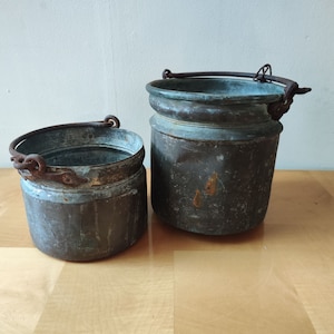 Puede incluir: Dos cubos de cobre antiguos con asas. El cubo más grande tiene una pátina verde desgastada y el cubo más pequeño tiene un acabado más pulido.