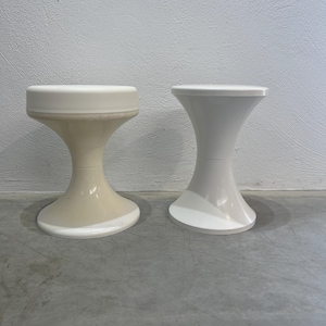 Può includere: Due sgabelli a forma di clessidra. Uno è bianco sporco, l'altro è bianco. Entrambi hanno una parte superiore e inferiore circolare, con un corpo curvo e svasato. Gli sgabelli sono realizzati in un materiale liscio e solido e sono appoggiati a una parete bianca.