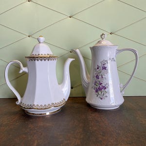 Può includere: Due teiere in porcellana bianca. Una è ottagonale con finiture dorate e coperchio abbinato. L'altra è più alta e sottile, con un motivo floreale viola e bianco. Entrambe hanno manici e beccucci curvi.