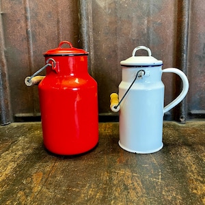 Può includere: Due lattine per il latte vintage in smalto. Una è rosso vivo con bordo nero e manico in legno. L'altra è bianca con bordo blu e manico bianco. Entrambe hanno coperchi e sono su una superficie di legno.