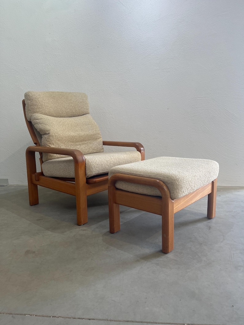 May include: A vintage armchair and footstool set. The chair features a high back with a cushioned headrest and armrests. The wooden frame and the cushions are a light beige colour. The footstool has a matching design.