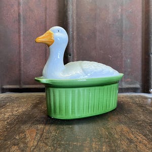 Könnte beinhalten: Eine keramische, gänseförmige Schale mit hellblauem Körper und leuchtend orangefarbenem Schnabel. Die Gans sitzt auf einem grünen, gerippten, ovalen Sockel. Die Schale dient wahrscheinlich zum Servieren oder Dekorieren.