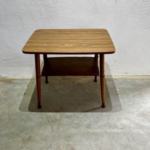 Może przedstawiać: Mały, kwadratowy drewniany stolik boczny z dolną półką. Blat i półka mają wzór słojów drewna. Stół ma cztery smukłe, zaokrąglone nogi. Stół ma około 51 cm szerokości i 51 cm długości.