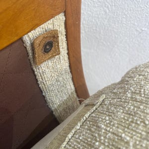 May include: Detail of a wooden chair showing a light brown wooden frame, beige fabric, and a brown leather button. The seat cushion is a light beige colour. The chair's design is visible.