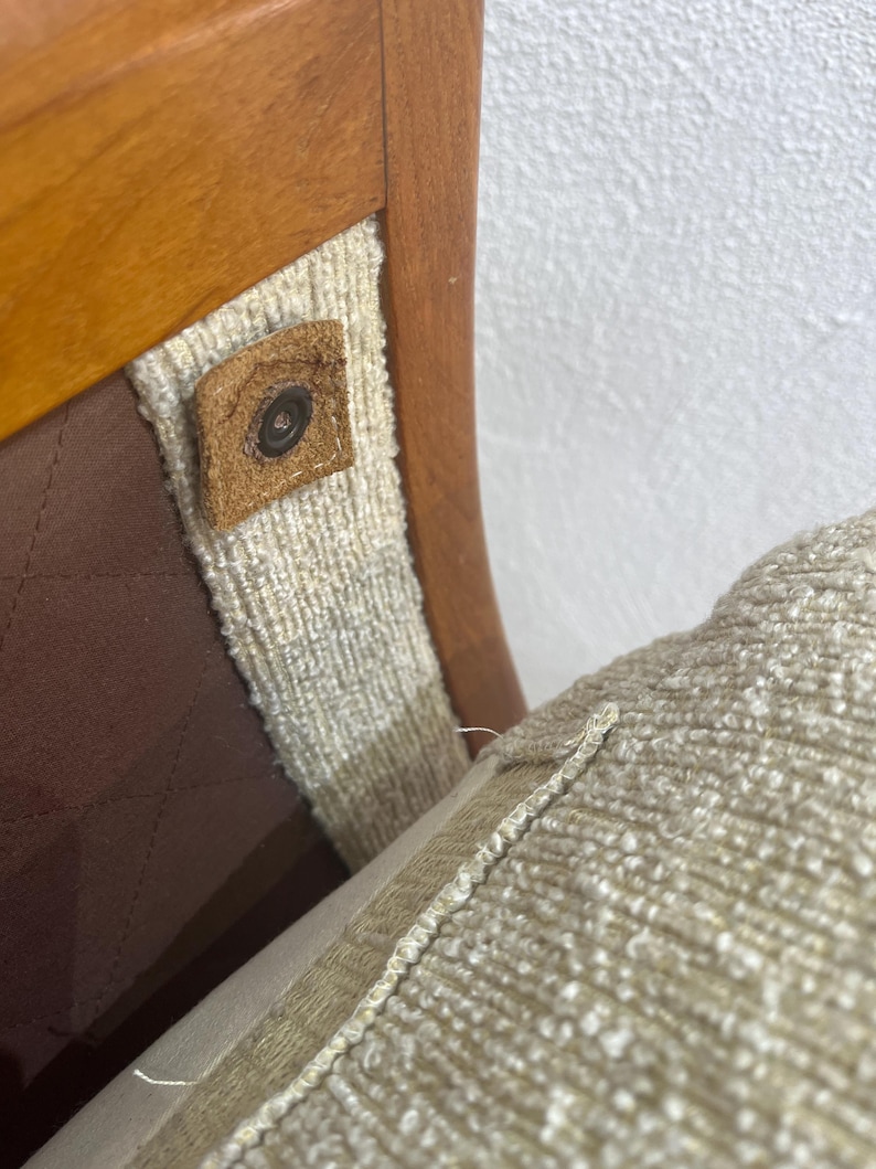 May include: Detail of a wooden chair showing a light brown wooden frame, beige fabric, and a brown leather button. The seat cushion is a light beige colour. The chair's design is visible.