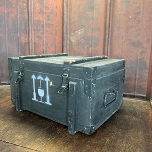 May include: A dark green wooden storage box with metal clasps and a handle. The box has a white painted symbol of arrows pointing upwards with a circle in the middle.