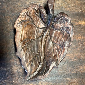 Könnte beinhalten: Ein handgefertigtes Tablett in Blattform aus Holz mit aufwendigen Schnitzereien. Das Tablett hat eine satte braune Farbe mit sichtbarer Holzmaserung und einer strukturierten Oberfläche. Die Adern des Blattes sind ebenfalls geschnitzt.
