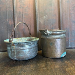 Könnte beinhalten: Zwei antike Kupferkessel mit gealterter Patina. Ein Topf hat einen Griff, der andere ist ein kleiner, zylindrischer Behälter. Das Kupfer hat stellenweise einen grünlich-blauen Farbton, der auf Alter und Gebrauch hindeutet. Die Töpfe stehen auf einer dunklen Holzoberfläche.