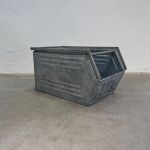 Op de afbeelding: Een rechthoekige opbergbak van gegalvaniseerd staal met een open voorzijde, schuine zijkanten en een horizontale stang aan de bovenzijde. De zijkant van de metalen bak is voorzien van de tekst Rapidbox en een klein labelhoudertje.
