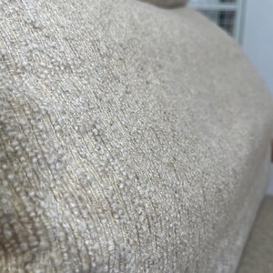 May include: Close-up of a beige fabric with a textured, woven appearance. The material has a nubby surface with variations in colour, creating a subtle, natural aesthetic. The fabric appears to be part of a larger piece of furniture.