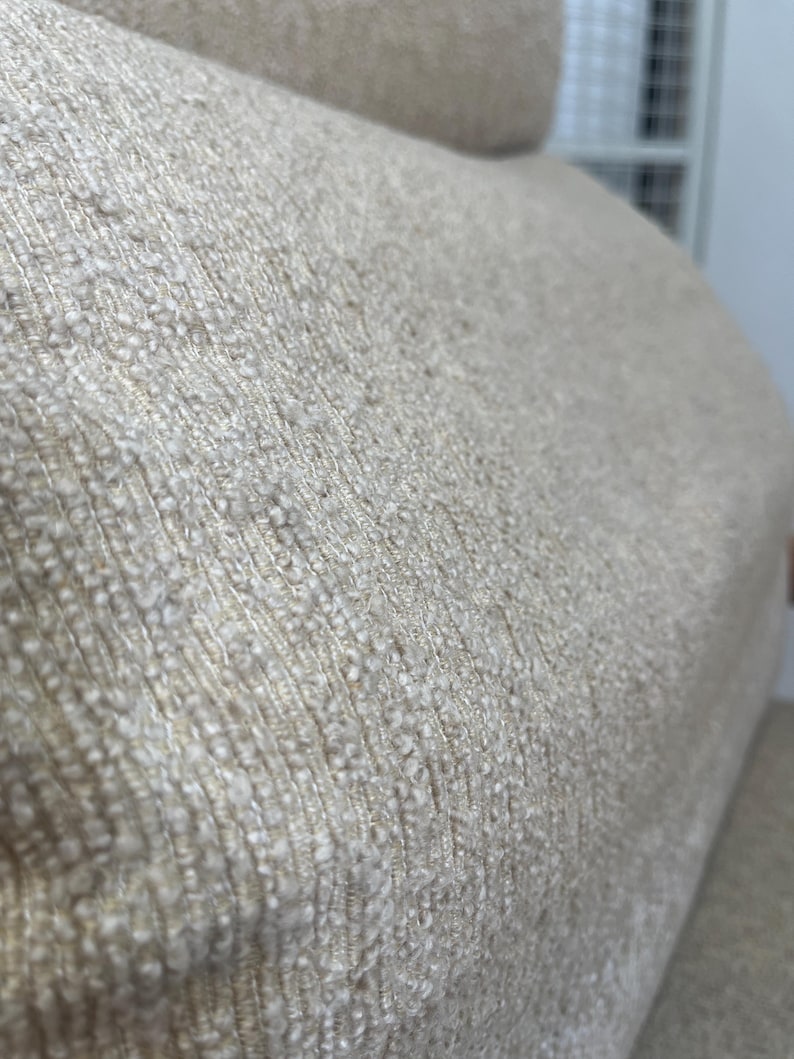 May include: Close-up of a beige fabric with a textured, woven appearance. The material has a nubby surface with variations in colour, creating a subtle, natural aesthetic. The fabric appears to be part of a larger piece of furniture.
