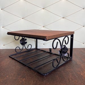 Puede incluir: Estantería de pared decorativa con una tapa de madera marrón oscuro y un marco de metal negro. El marco presenta volutas y detalles florales. La estantería está diseñada para montarse en la pared y tiene forma rectangular.