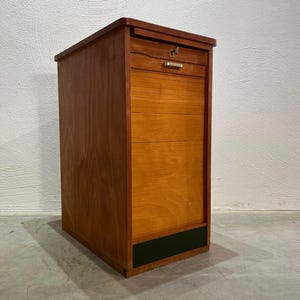 Könnte beinhalten: Ein Holzschrank mit Rolltür und Schublade. Der Schrank besteht aus hellem und dunklem braunem Holz mit einem schwarzen Streifen am Boden. Die Schublade hat einen kleinen, goldfarbenen Griff. Der Schrank ist rechteckig und steht auf einem Betonboden.