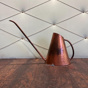 May include: A copper watering can with a long, slender spout and a hammered texture. The can has a conical body and a curved handle. The copper has a rich, reddish-brown color, and the spout extends to the left.