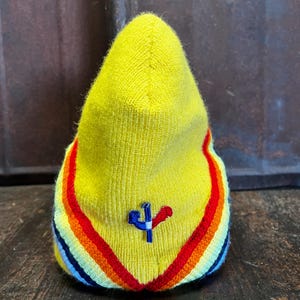 Op de afbeelding: Een gele gebreide muts met een regenboogstreepontwerp aan de basis. De muts heeft een klein geborduurd embleem in blauw en rood. De muts is gemaakt van een zacht, getextureerd materiaal.