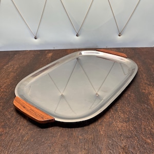 Può includere: Un vassoio da portata argentato con due manici in legno. Il vassoio è rettangolare e ha un bordo leggermente arrotondato. I manici in legno sono di colore marrone scuro.