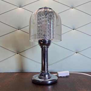 Op de afbeelding: Een chromen tafellamp met een heldere, klokvormige glazen kap. De kap omsluit een getextureerde, cilindrische lichtbron. De lamp heeft een ronde voet en een cilindrische steel. Een wit netsnoer loopt vanaf de basis.