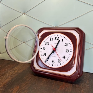 Op de afbeelding: Een vierkante, bruin omrande quartz klok met een witte wijzerplaat en zwarte cijfers. De klok heeft zwarte wijzers en een rode secondewijzer. Een vergrootglas is voor de klok geplaatst.