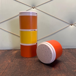 Può includere: Una pila di tre contenitori cilindrici colorati in arancione, giallo e arancione, con fasce bianche. Un contenitore è sdraiato su un lato, mostrando un coperchio perforato. I contenitori sono su una superficie di legno scuro.