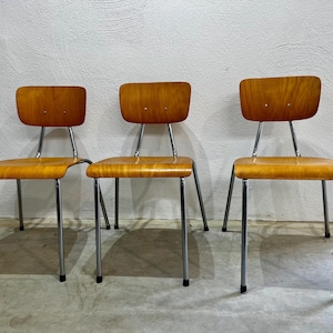 Op de afbeelding: Drie mid-century moderne stoelen met gebogen houten zittingen en rugleuningen. De stoelen hebben een lichtbruine afwerking en chromen poten. De stoelen zijn in een rij opgesteld tegen een witte muur.