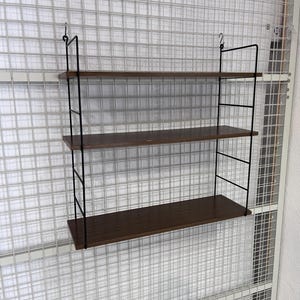 Könnte beinhalten: Ein Wandregal mit drei Holzböden und einem schwarzen Metallrahmen. Die Regalböden sind aus dunklem Holz und der Rahmen besteht aus dünnen Metallstäben.