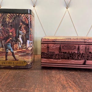 Peut inclure: Deux boîtes en étain vintage. La première boîte est verte et présente une illustration colorée de personnes dans un cadre tropical. Le texte sur la boîte indique "Jacobs Kaffee". La deuxième boîte est rose et présente une illustration en noir et blanc d'un horizon urbain. Le texte sur la boîte indique "Bremen 1568".