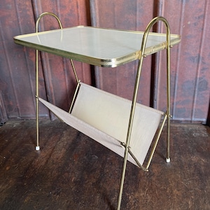 Puede incluir: Una mesa auxiliar vintage con tapa rectangular de cristal y estructura metálica dorada. La mesa tiene un revistero incorporado de tela beige. Las patas tienen topes blancos. Una pieza de acento elegante.