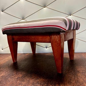 Könnte beinhalten: Ein kleiner Fußhocker aus Holz mit einem rechteckigen, gestreiften Kissen. Das Kissen hat ein Muster aus grauen, schwarzen und roten Streifen. Die Beine des Hockers sind rotbraun.