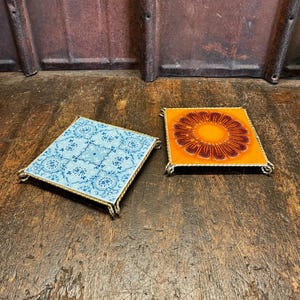 Könnte beinhalten: Zwei quadratische Keramikuntersetzer. Einer ist hellblau mit einem Blumenmuster, der andere orange mit einem braunen Sonnenmuster. Beide haben goldfarbene Metallrahmen und werden auf einer Holzoberfläche präsentiert.