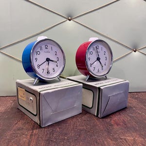 May include: Two vintage alarm clocks, one blue and one red, with white faces and silver legs. The clocks sit on their original gray boxes. The clock faces display the brand name "ruhle". The background is a light green wall with a diamond pattern.