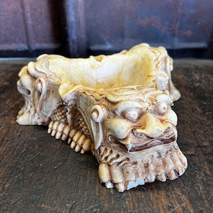 Puede incluir: Un cenicero decorativo beige y marrón. El cenicero está esculpido en forma de criatura mítica con una cara detallada, garras y escamas. El cenicero tiene forma rectangular con un cuenco poco profundo para contener las cenizas.