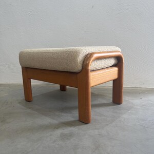 May include: A mid-century style footstool featuring a light beige upholstered cushion and a solid wood frame. The frame is a warm, natural wood tone, and the stool has a rectangular shape with rounded edges. The legs are short and sturdy.