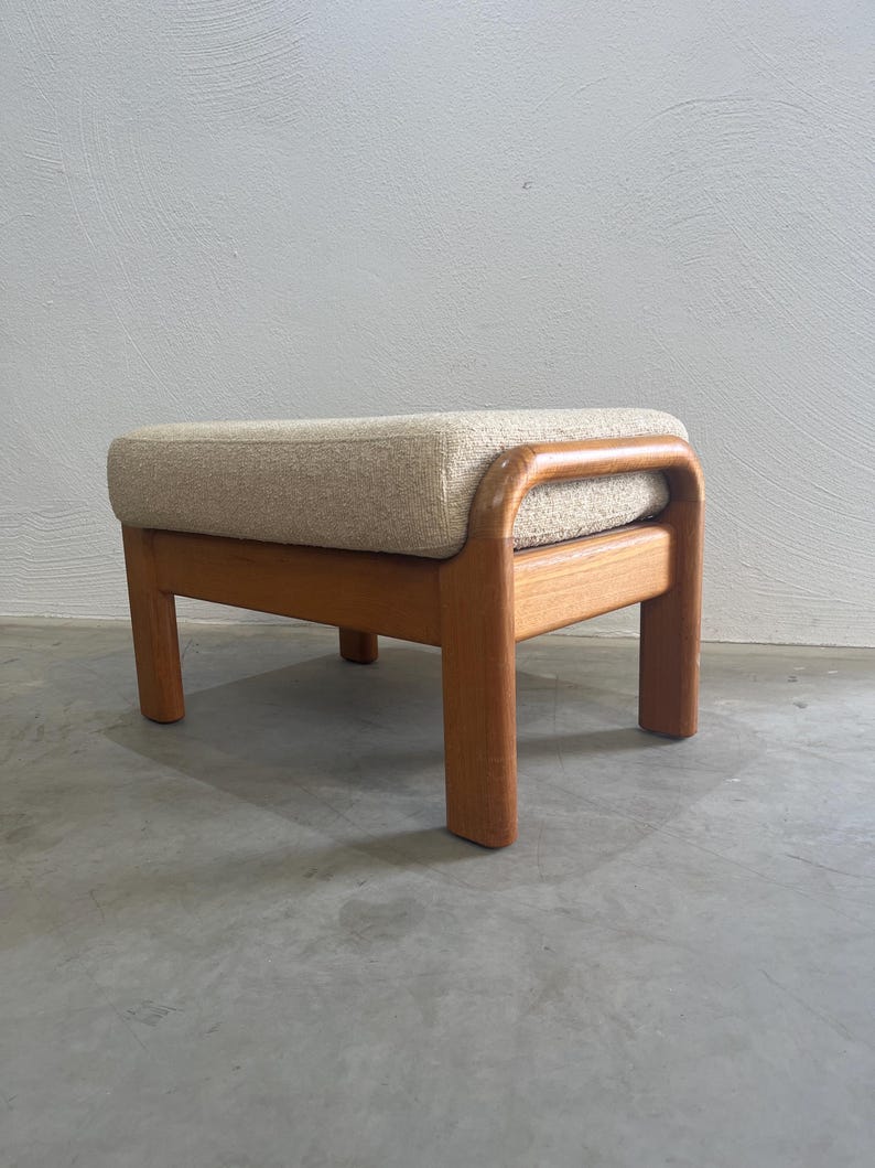 May include: A mid-century style footstool featuring a light beige upholstered cushion and a solid wood frame. The frame is a warm, natural wood tone, and the stool has a rectangular shape with rounded edges. The legs are short and sturdy.