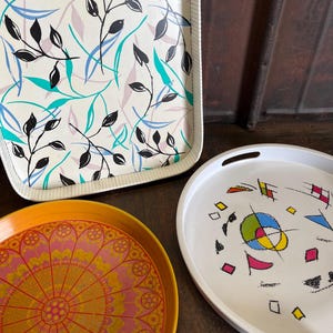 May include: Three decorative trays with different patterns. The first tray is rectangular and has a white background with black and teal leaf designs. The second tray is round and has a white background with a colourful abstract design. The third tray is round and has an orange background with a pink and yellow mandala design.