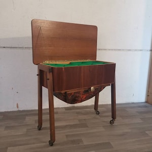 Puede incluir: Un gabinete de costura de madera vintage con una tapa abatible y un cajón extraíble. El gabinete tiene un interior forrado de fieltro verde y una cesta forrada de tela estampada. El gabinete está sobre ruedas y tiene un acabado de madera oscura.