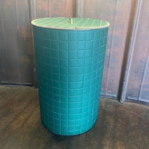 Può includere: Un contenitore cilindrico color verde acqua con un motivo a griglia quadrata. Il coperchio è anch'esso verde acqua con un motivo a griglia e un bordo dorato. Il contenitore è su una superficie di legno scuro.